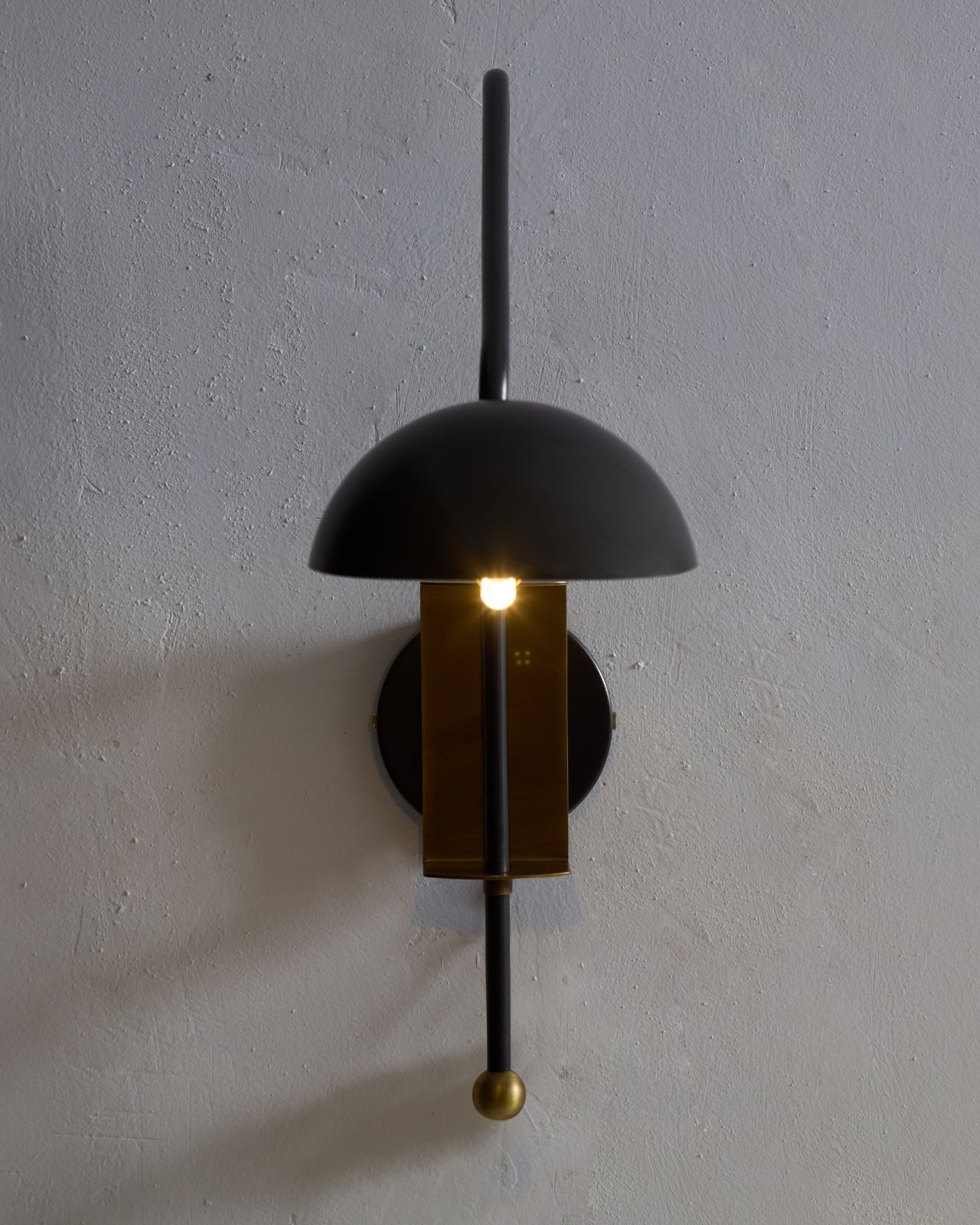 Marcello wall sconce mounted on a textured wall, featuring a black dome shade, vertical black stem, and brass center with warm illuminated bulb.