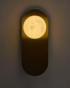 Milan wall sconce lit on a textured wall, featuring a brushed brass vertical backplate and a glowing round white glass globe that casts a warm ambient light.