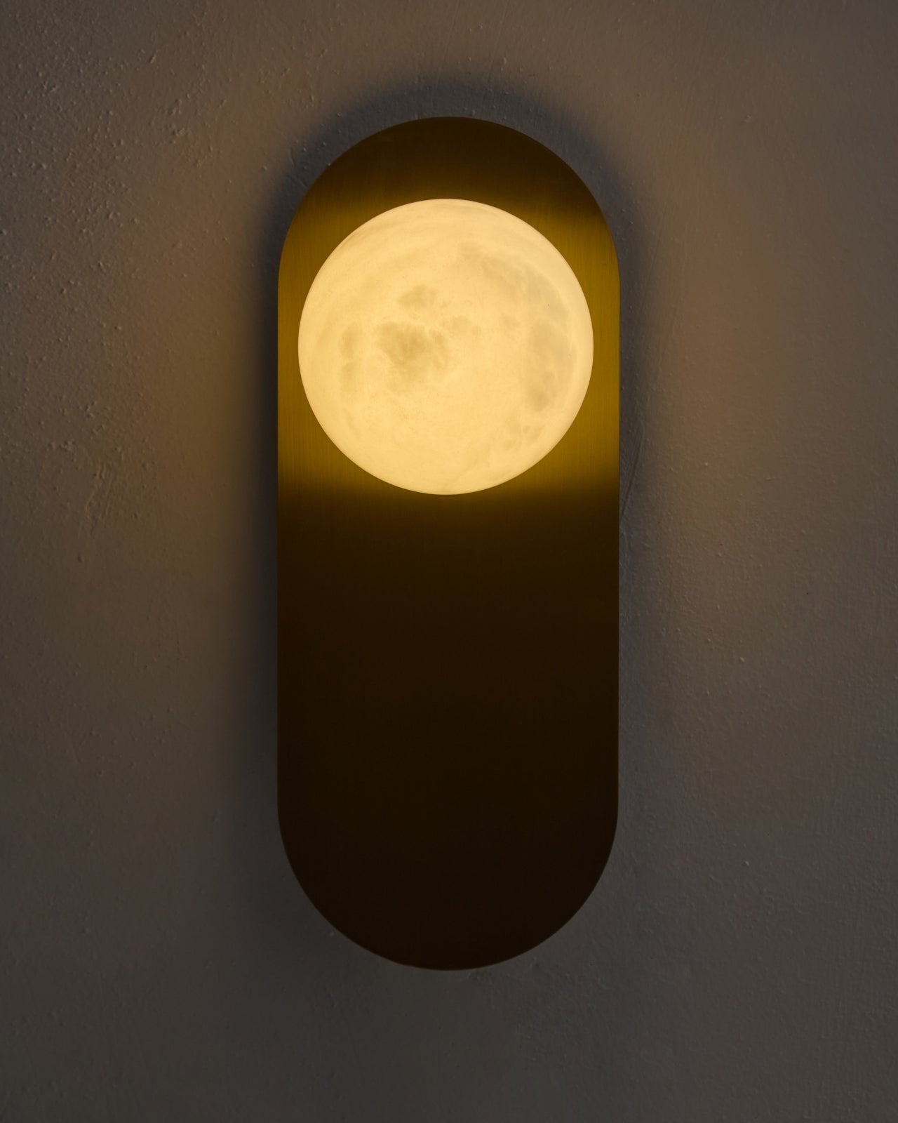 Milan wall sconce lit on a textured wall, featuring a brushed brass vertical backplate and a glowing round white glass globe that casts a warm ambient light.