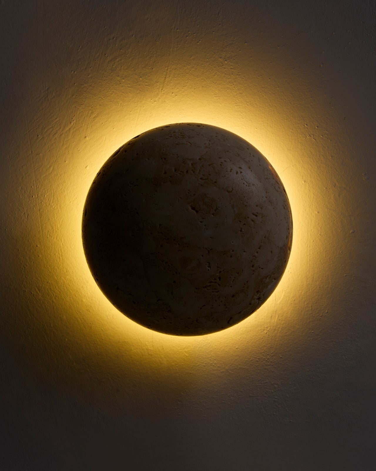 Wall-mounted spherical travertine sconce, illuminated and casting a warm halo glow on the wall.