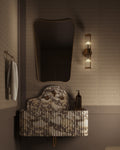 Butterfly Mirror with an irregular, wing-inspired shape mounted above a wall-mounted sink, styled with a vertical wall light, towel hook, and minimal accessories in a softly lit bathroom.