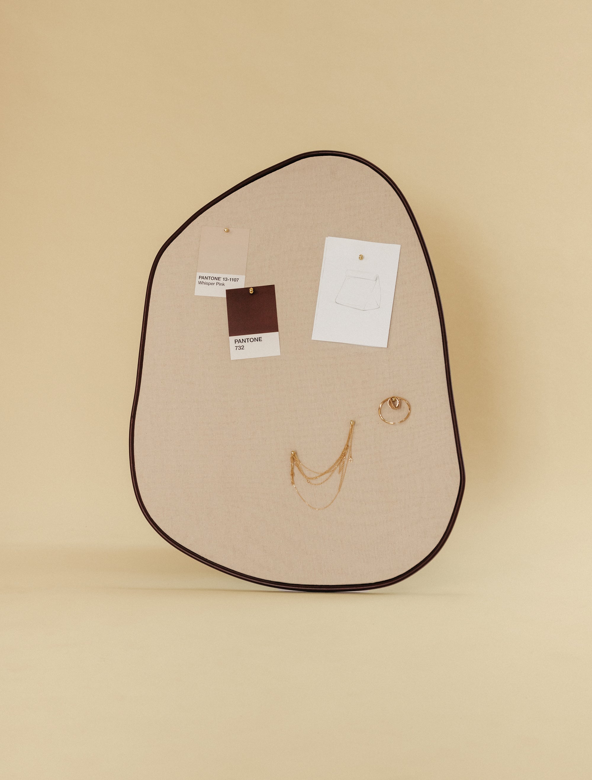 Asymmetrical fabric-covered pinboard with a brown outer frame, featuring pinned Pantone color cards, a small sketch, and gold jewelry.
