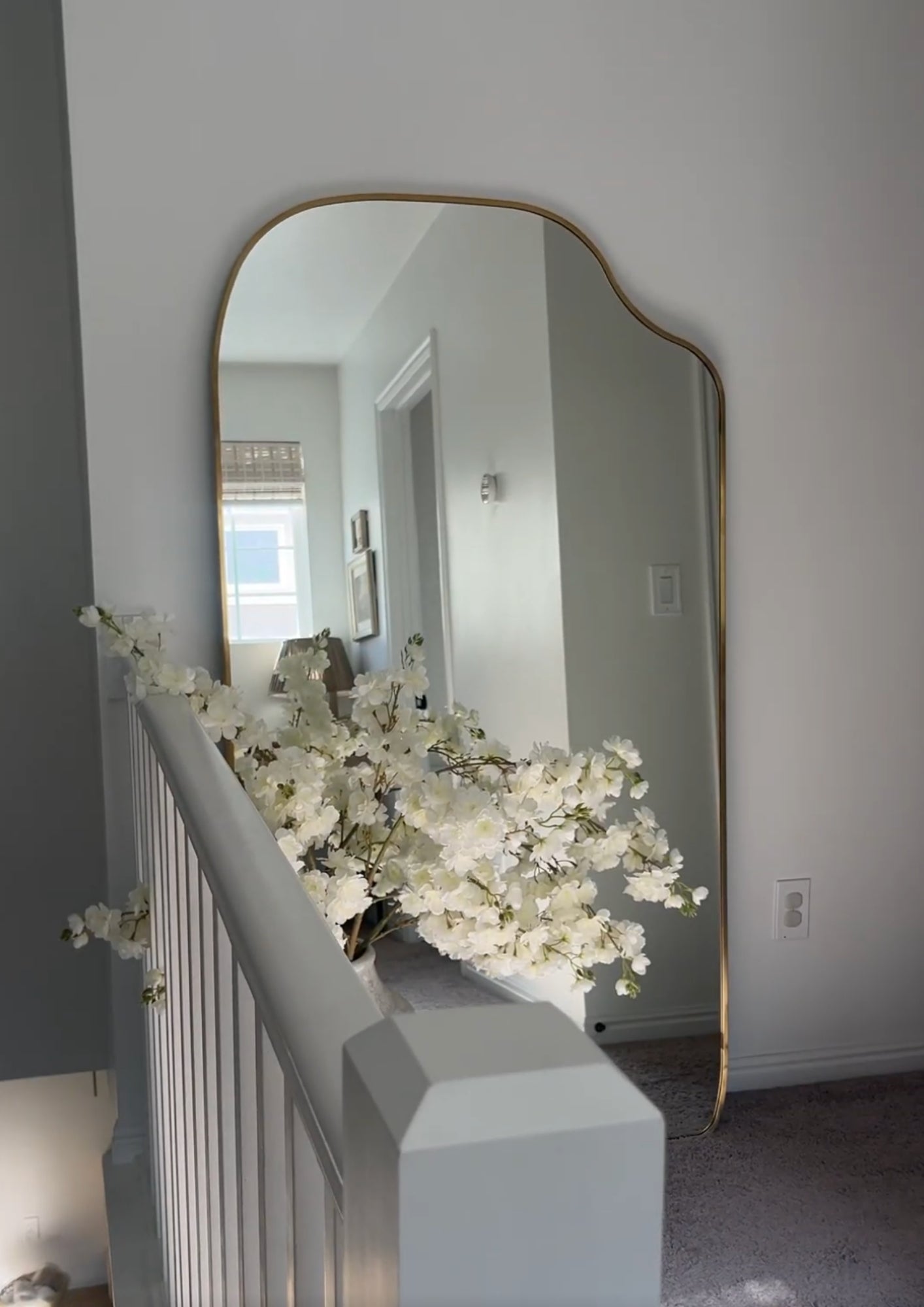 Organic-shaped full length mirror with brass frame, placed near a staircase and styled with white flowers.