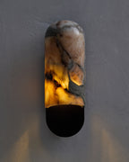 Marble wall sconce mounted on a textured gray wall, featuring a rounded vertical form with natural beige, gray, and amber veining, softly illuminated from within above a gunblack metal base.