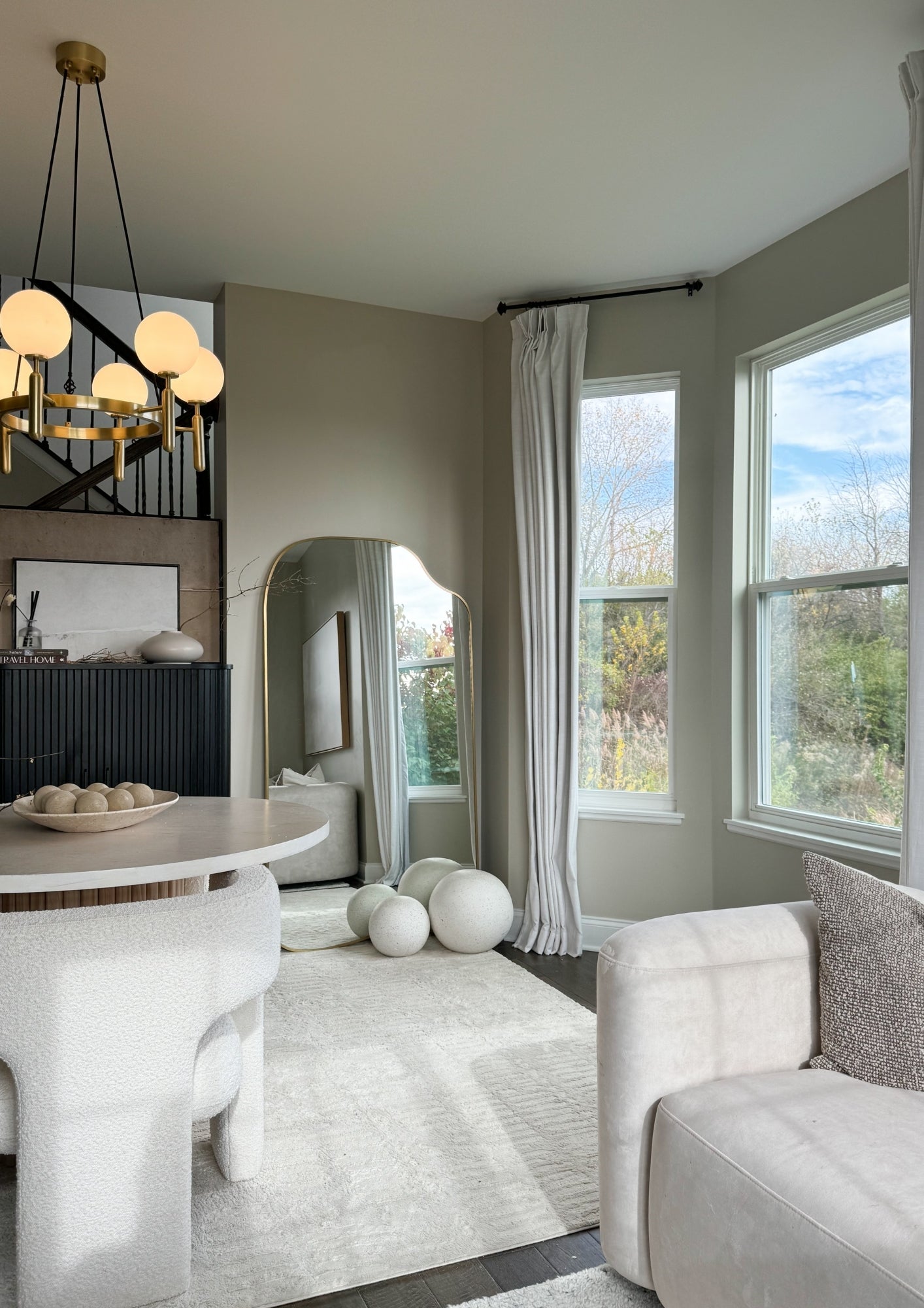 Organic-shaped full length mirror with brass frame in a bright living room with large windows and neutral decor.