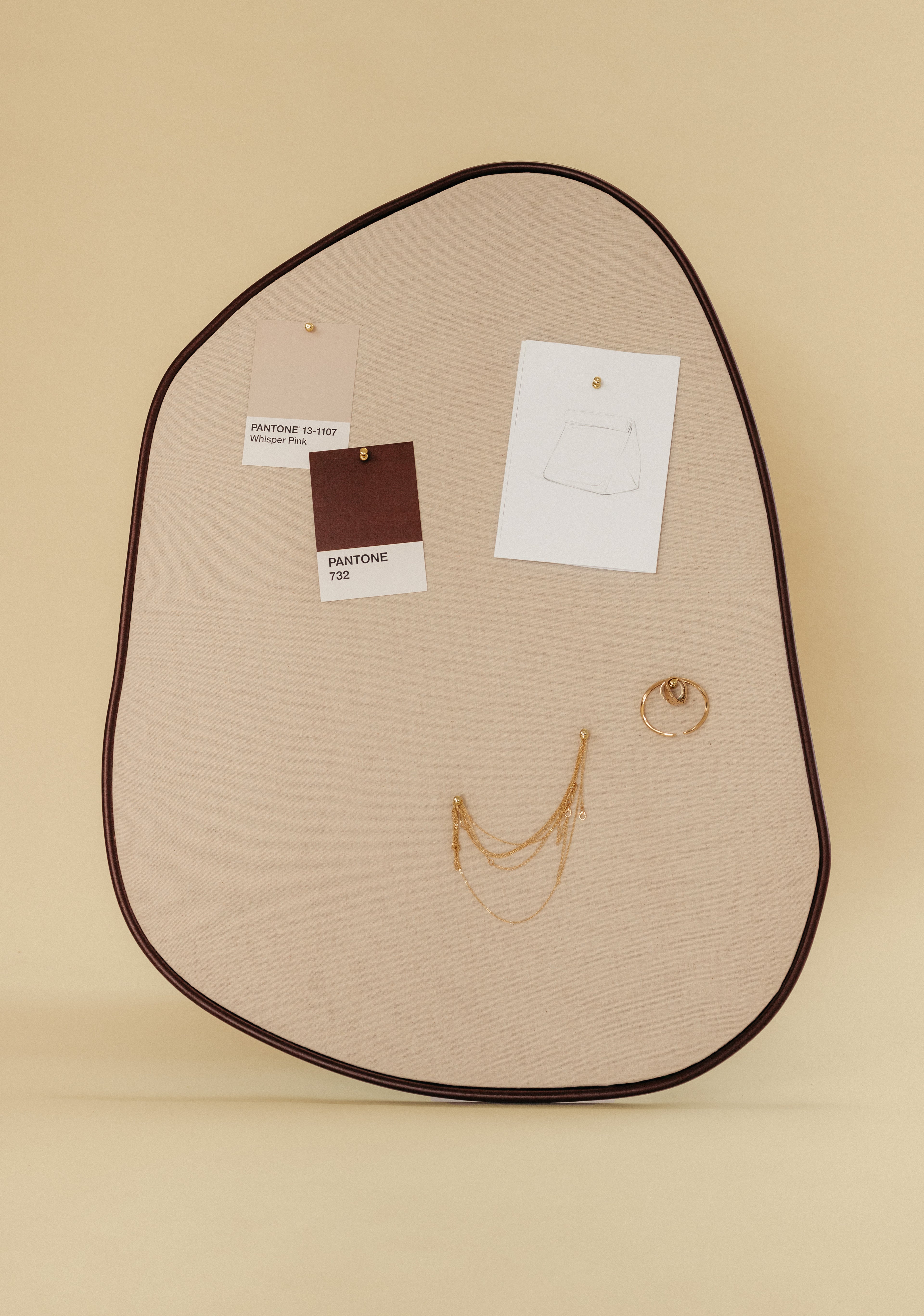 Asymmetrical beige fabric pinboard with a brown outer edge, featuring pinned Pantone color cards, a paper sketch, and hanging gold jewelry on a light background.