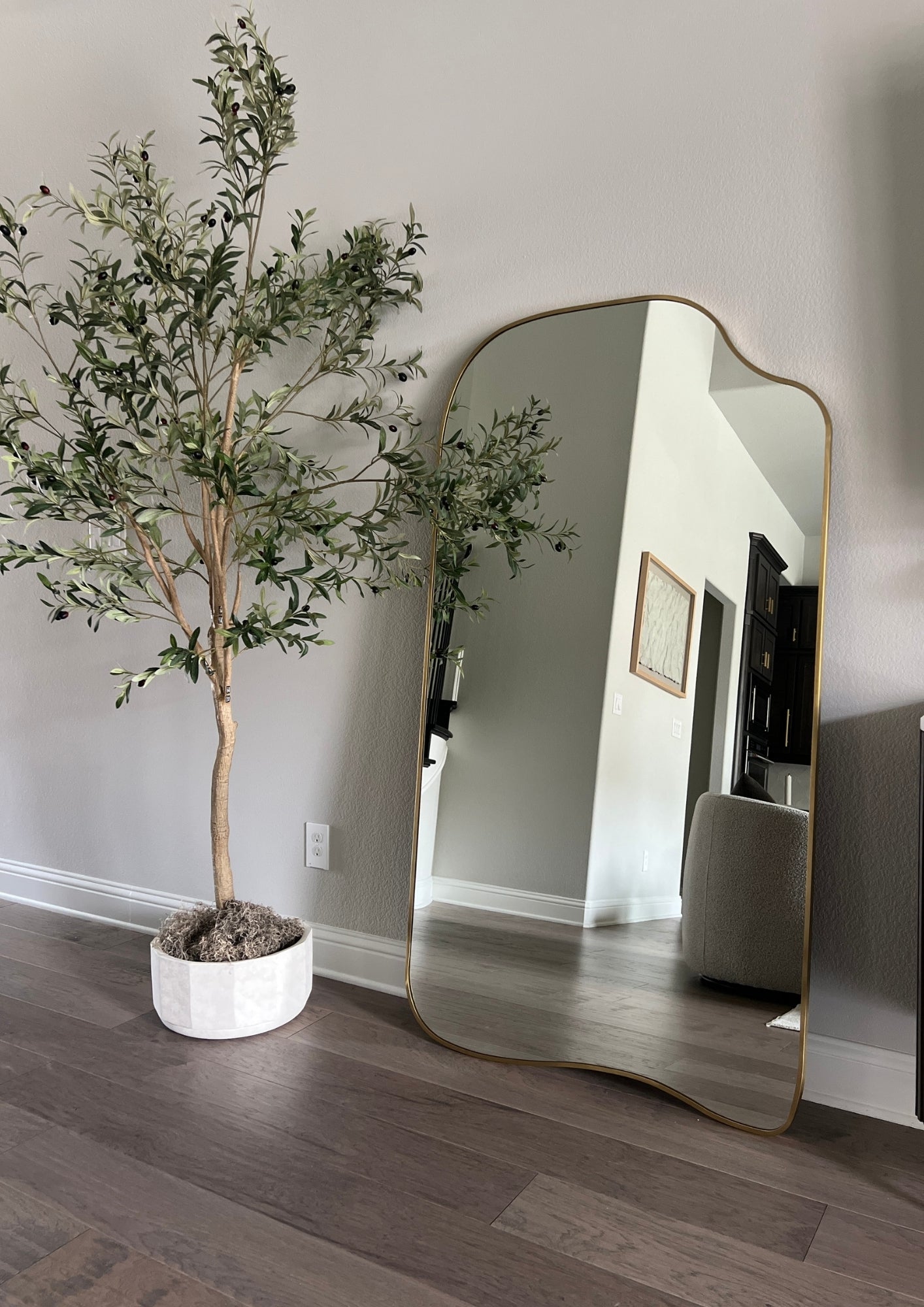 Organic-shaped full length mirror with brass frame, placed beside a potted tree in a minimalist interior.