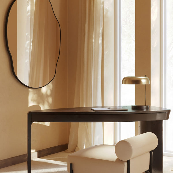 Irregular-shaped wall mirror with a slim dark frame mounted above a modern desk, reflecting sheer curtains and natural daylight.