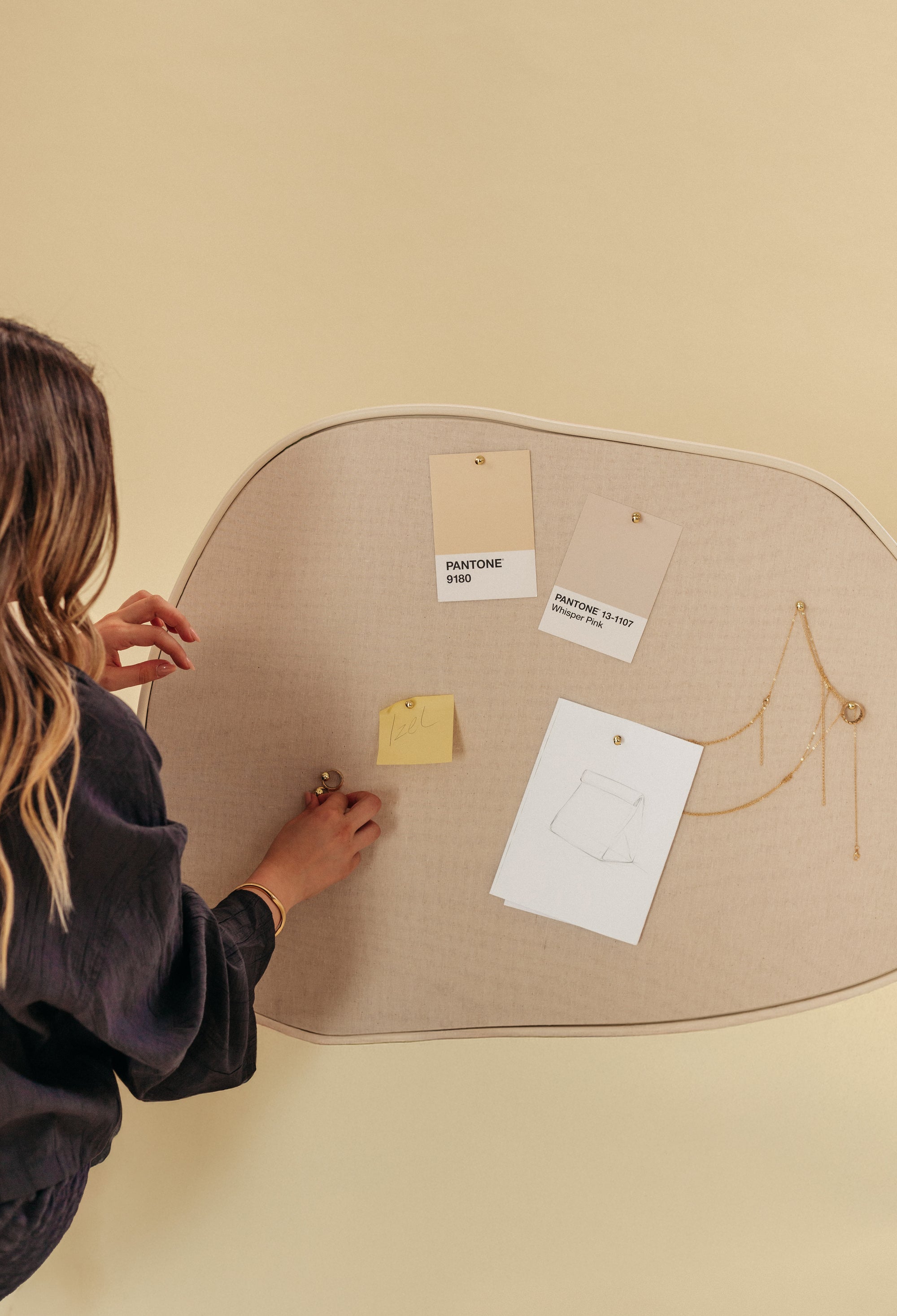 Asymmetrical beige fabric pinboard shown with hands pinning jewelry, Pantone color cards, and a paper sketch on a soft neutral background.