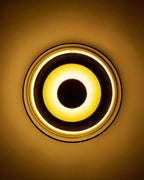Close-up view of a wall-mounted brass sconce with a circular design, showing an illuminated ring of light surrounding a central dark circular element against a wall.