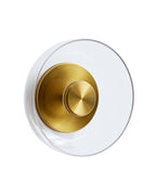 Front-facing view of a wall-mounted sconce featuring a circular brass center with a clear glass disc surrounding it, shown against a white background.