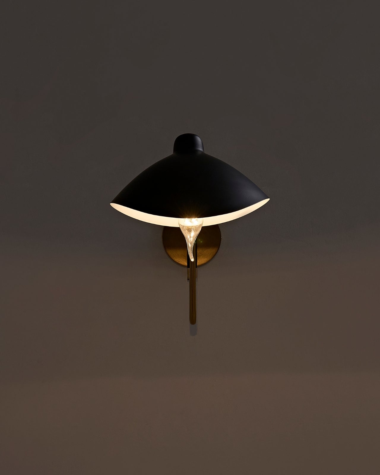 Wall-mounted sconce illuminated on a dark wall, featuring a black dome-shaped metal shade with a white interior, a brass wall plate and vertical stem, and an exposed bulb casting a warm downward glow.