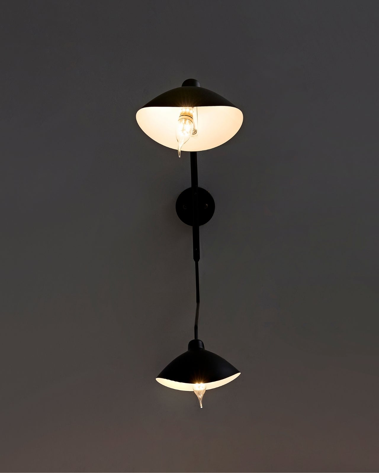 Fleur de Noir Wall Sconce illuminated on a dark wall, featuring a matte black vertical stem with two opposing dome-shaped shades casting a warm glow.