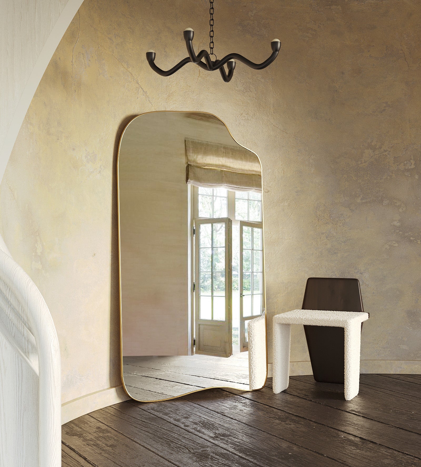 Asymmetrical full-length mirror with an organic shape and thin brass-colored frame, leaning against a textured wall in a softly lit room with wood flooring and sculptural furniture.