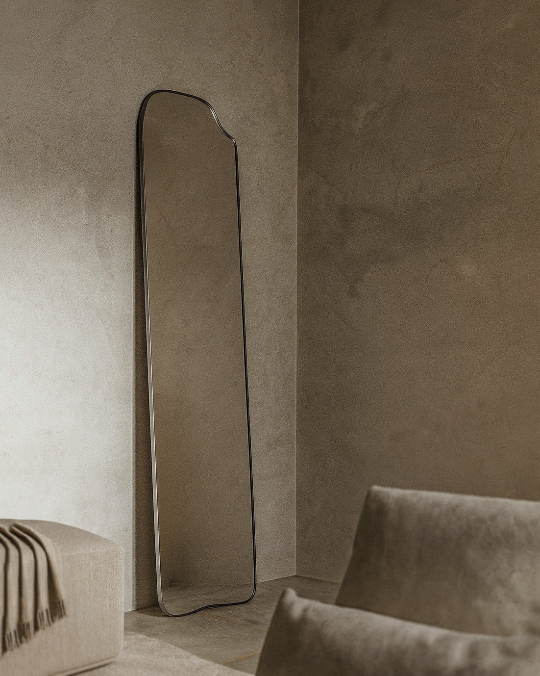 Asymmetrical full-length mirror with an organic shape and thin dark frame, leaning against a textured wall in a living room with soft natural light and neutral upholstered seating nearby.