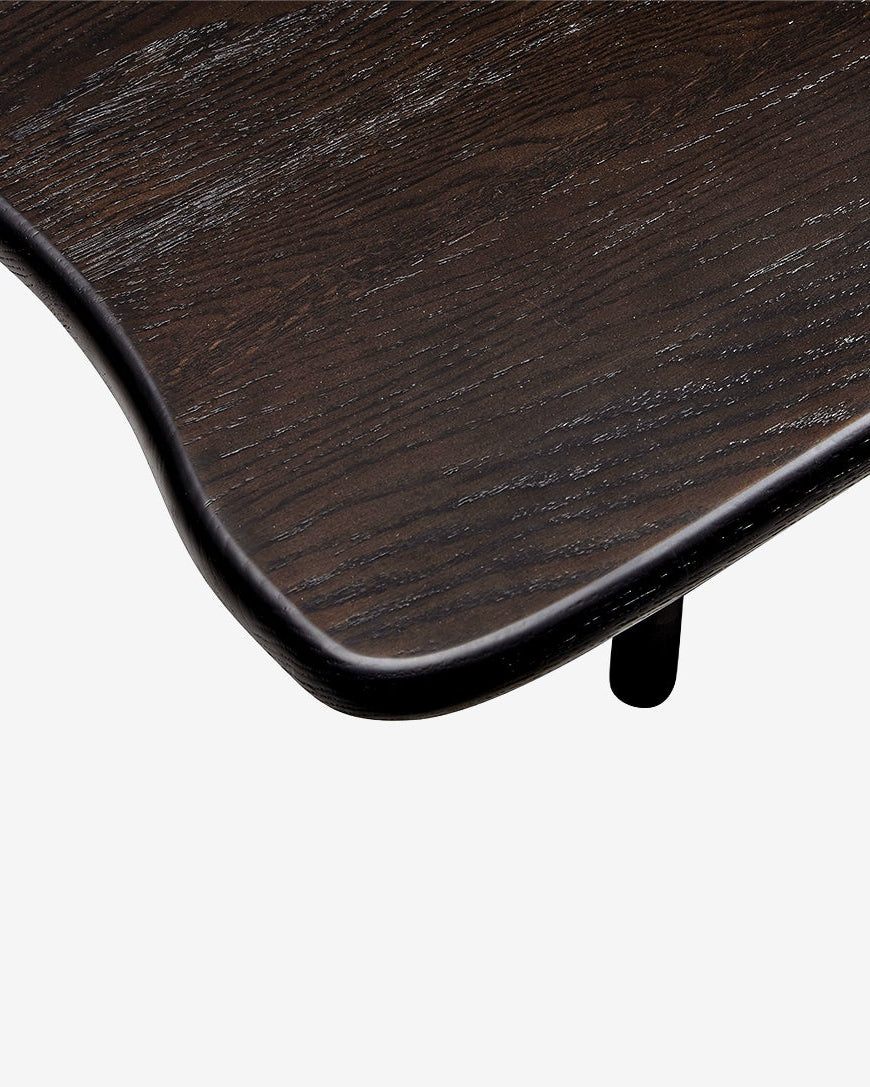 Close-up detail of a dark brown wooden table corner with a smooth curved edge and a small black leg, shown against a light gray background.