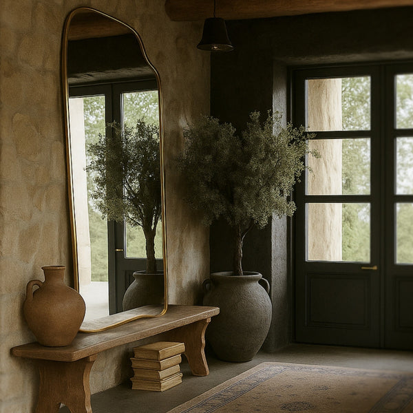 Asymmetrical full-length mirror with an organic shape and thin brass-colored frame, mounted above a wooden bench in a rustic entryway with potted plants and natural light.
