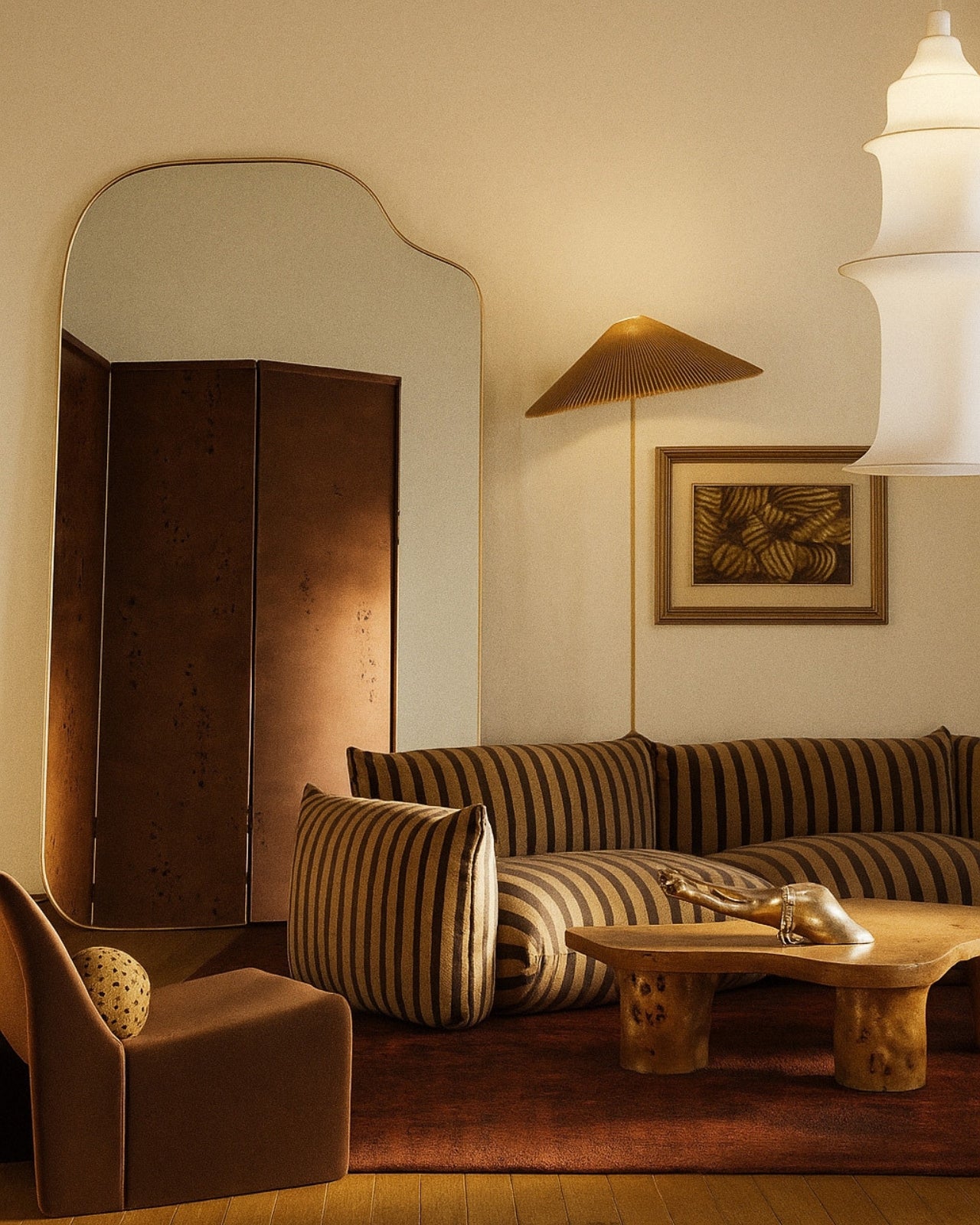 Asymmetrical full-length mirror with an organic shape and thin brass-colored frame, styled in a warm living room with striped seating, sculptural furniture, and ambient lighting.