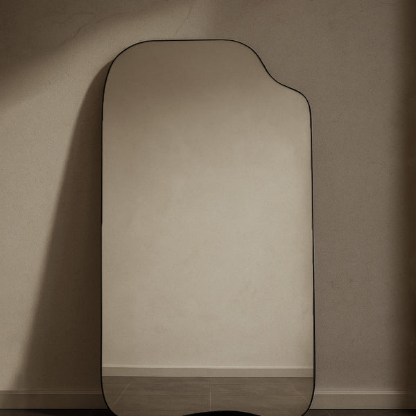 Asymmetrical full-length mirror with an organic shape and thin black frame, shown standing against a neutral wall on the floor.