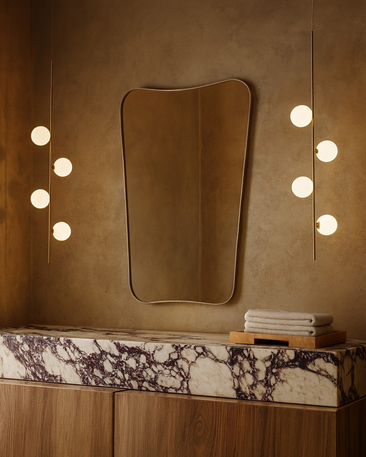 Warm modern bathroom with an irregular wall mirror, marble countertop, wood vanity, stacked towels, and vertical globe sconces.