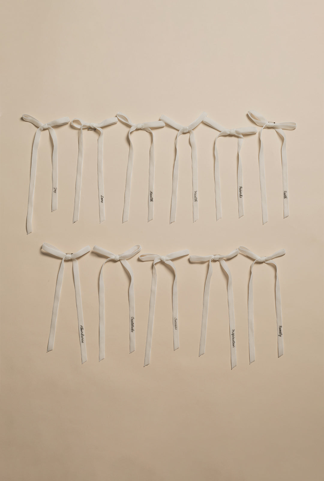 Eleven white velvet ribbon bows laid out in two rows on a neutral background, each ribbon embroidered with a single word representing a wish, such as joy, love, health, and gratitude.