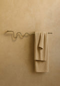 Wall-mounted towel bar with a wavy brass-colored design, holding a folded beige towel against a textured neutral wall.