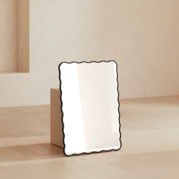 Rectangular wall mirror with a black frame and softly wavy edges, leaning against a wall in a neutral interior setting.