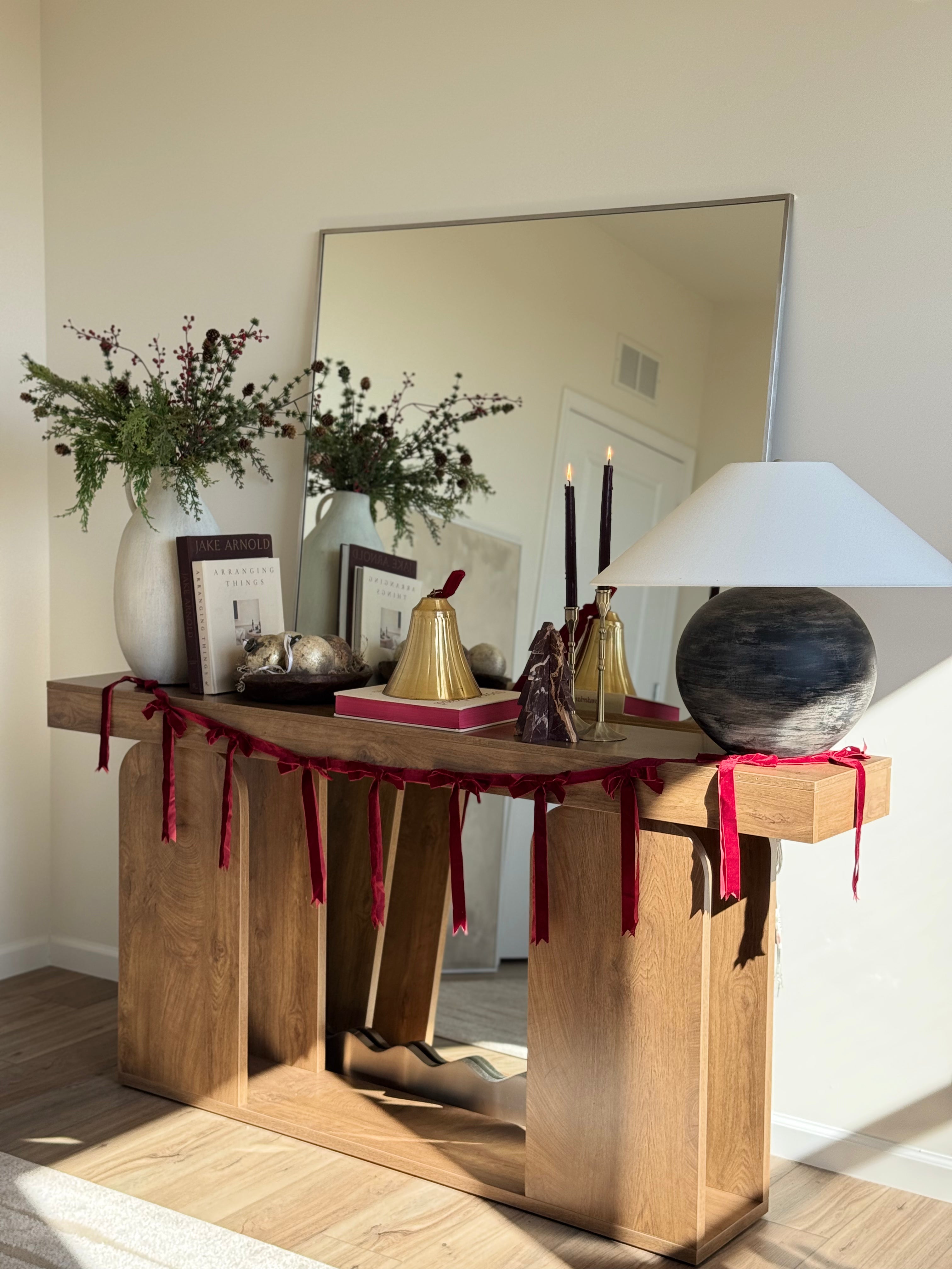 A full-length rectangular mirror is placed behind a wooden console table, styled with holiday greenery, candles, books, and red ribbon accents in a softly sunlit interior.