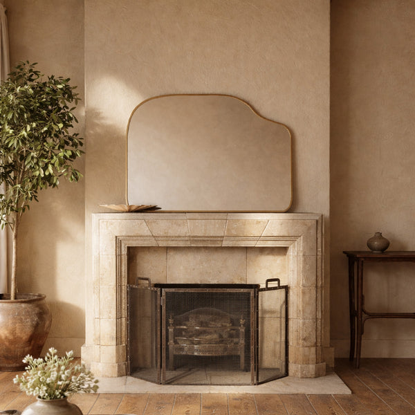 Asymmetrical brass-framed mirror mounted above a stone fireplace in a neutral interior setting.