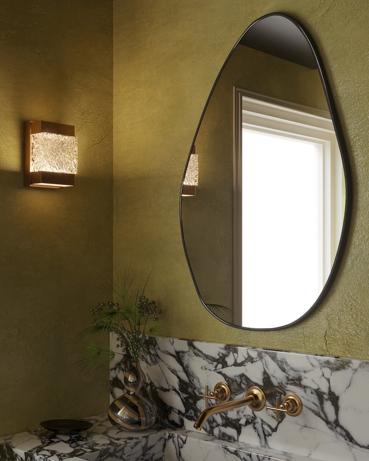 Frameless pebble-shaped bathroom mirror with an organic silhouette, styled above a marble vanity with brass fixtures and textured green walls.