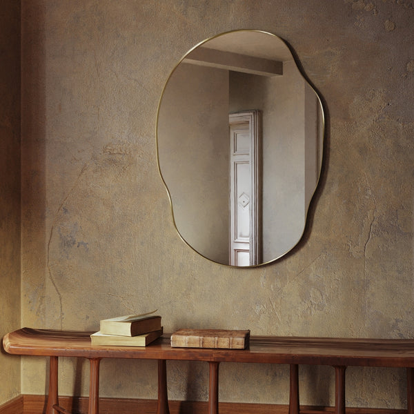 Irregular-shaped wall mirror with a brass frame mounted on a textured wall above a wooden bench with stacked books.