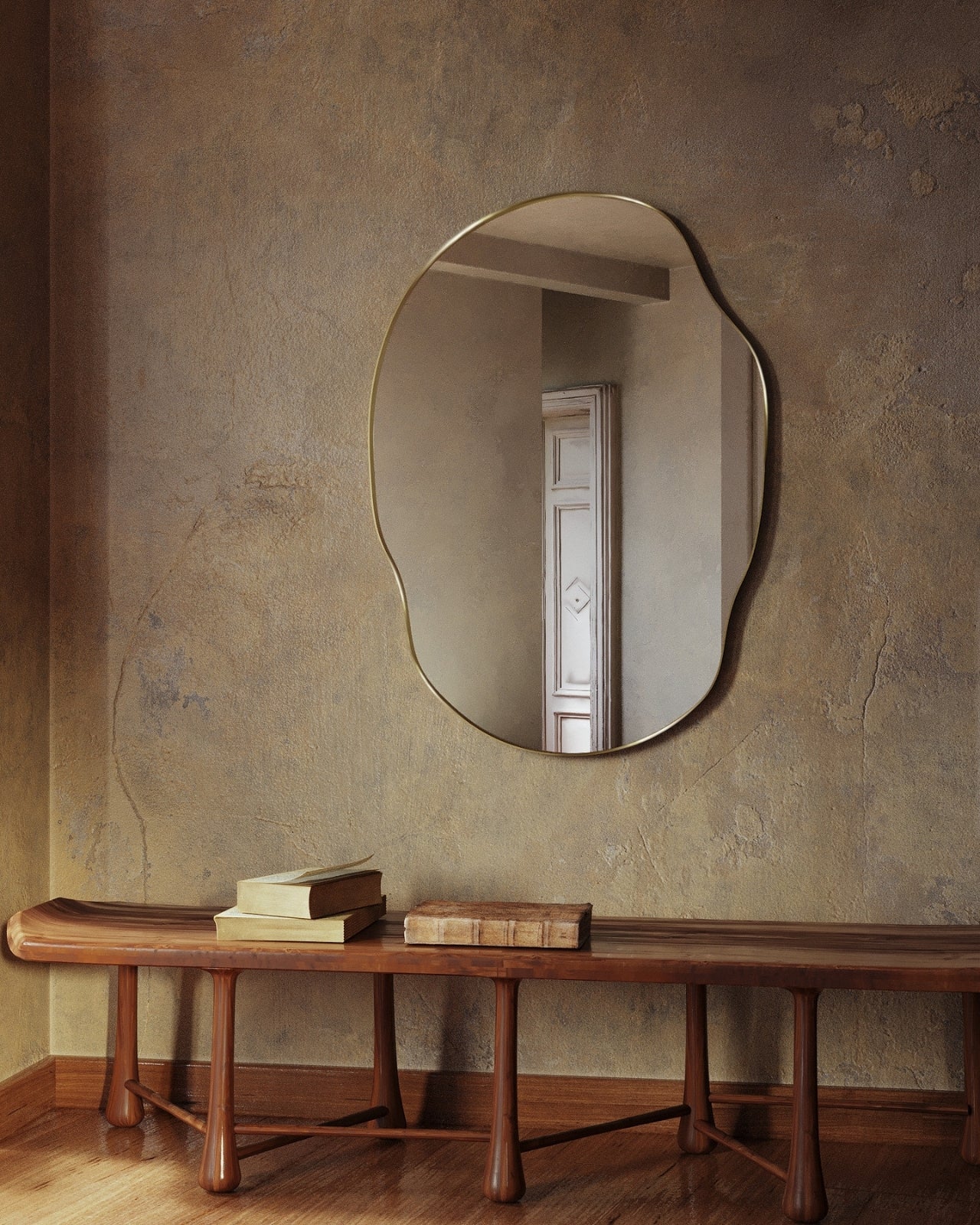 Irregular-shaped wall mirror with a brass frame mounted on a textured wall above a wooden bench with stacked books.