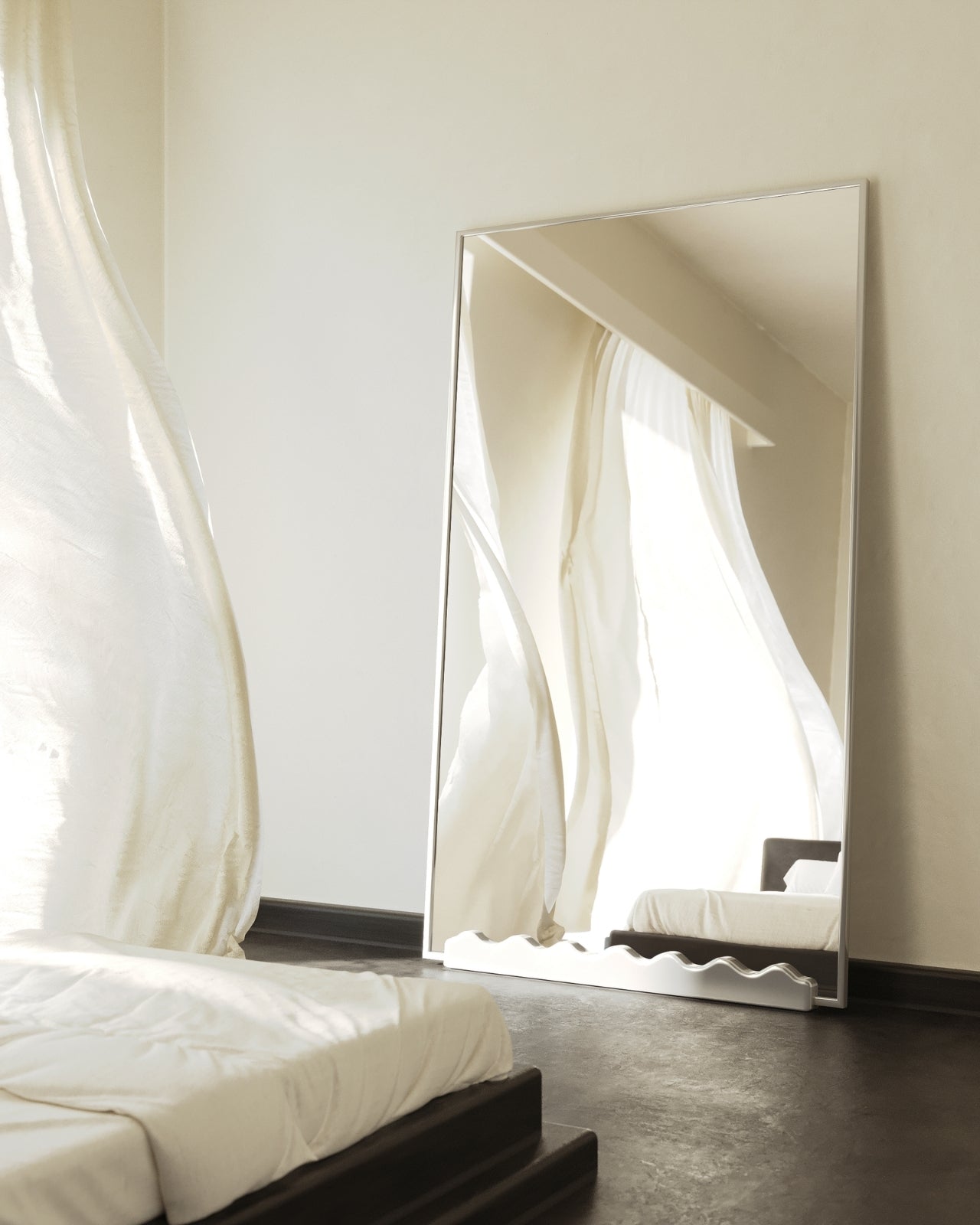 Full-length rectangular mirror with stainless steel frame leaning against a bedroom wall, reflecting sheer curtains and a bed, with a decorative wavy detail at the base.
