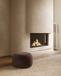 Pebble-style round dark brown ottoman placed on a rug beside a fireplace.