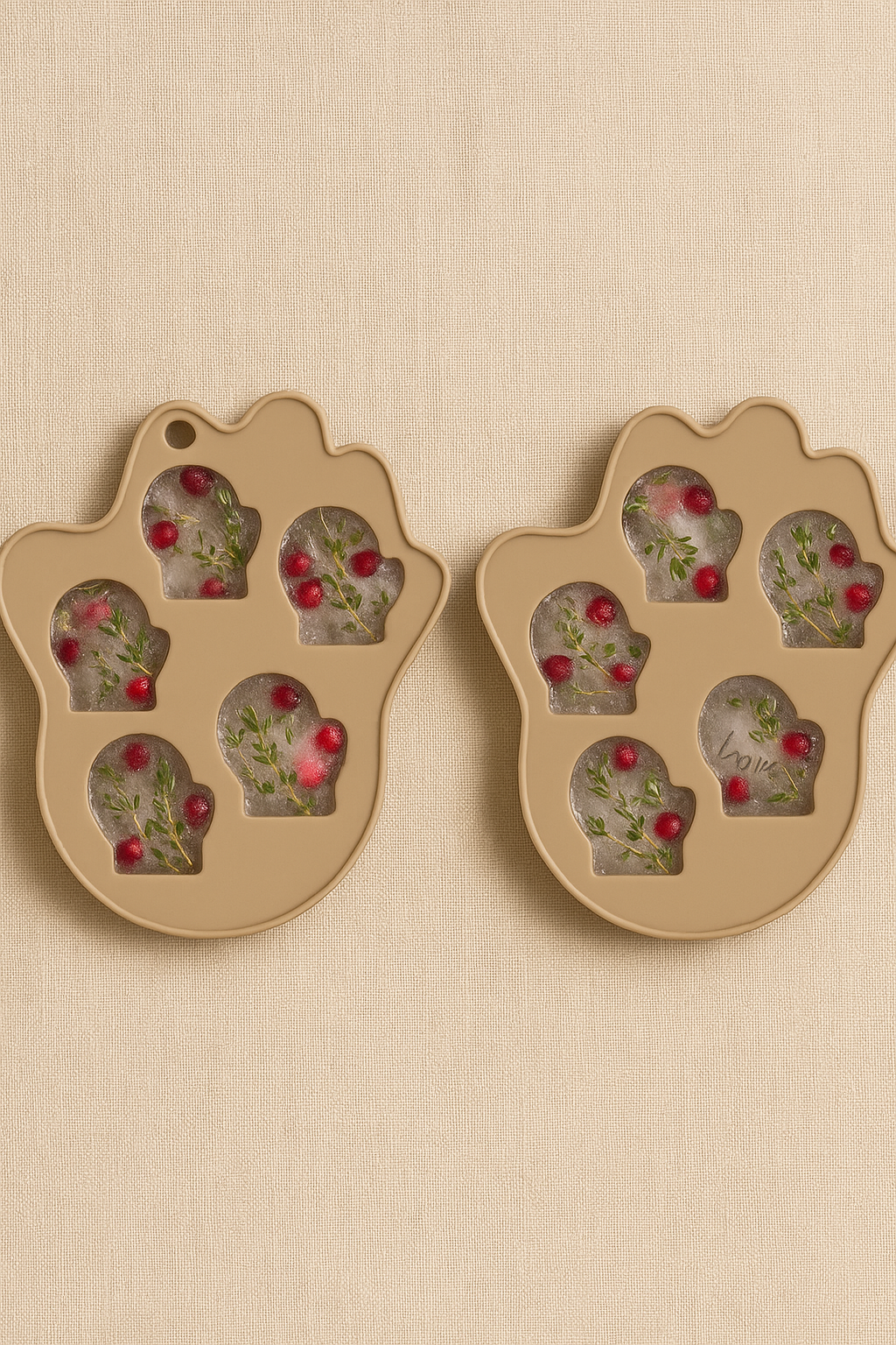 Two beige mitten-shaped silicone kitchen molds placed side by side, each filled with frozen liquid containing pomegranate seeds and rosemary sprigs.