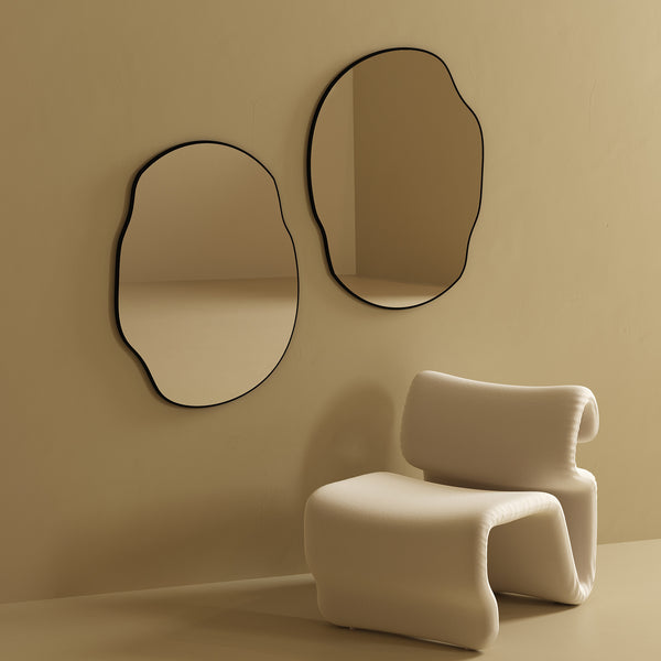 Two irregular-shaped wall mirrors with black frames mounted side by side on a beige wall above a sculptural upholstered chair.