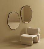 Two irregular-shaped wall mirrors with black frames mounted side by side on a beige wall above a sculptural upholstered chair.
