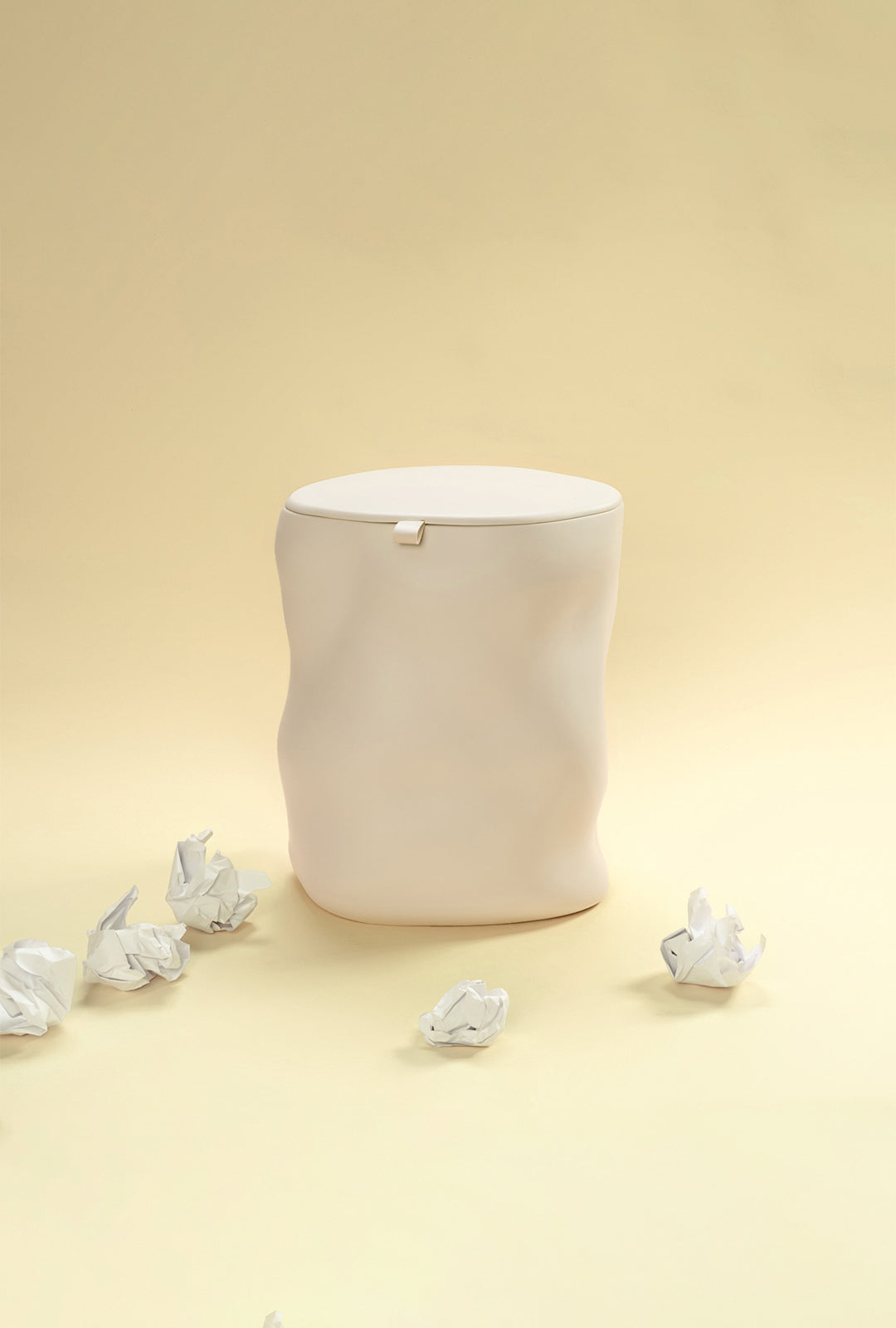 A beige sculptural dustbin with a fitted lid, surrounded by several crumpled pieces of white paper on the floor, against a light yellow background.