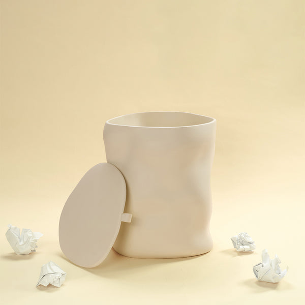 A beige sculptural dustbin shown with its lid removed and placed to the side, with several crumpled pieces of white paper on the floor around it, against a light yellow background.