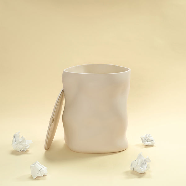 A beige sculptural dustbin shown open with its lid leaning against the side, surrounded by several crumpled pieces of white paper, against a light yellow background.