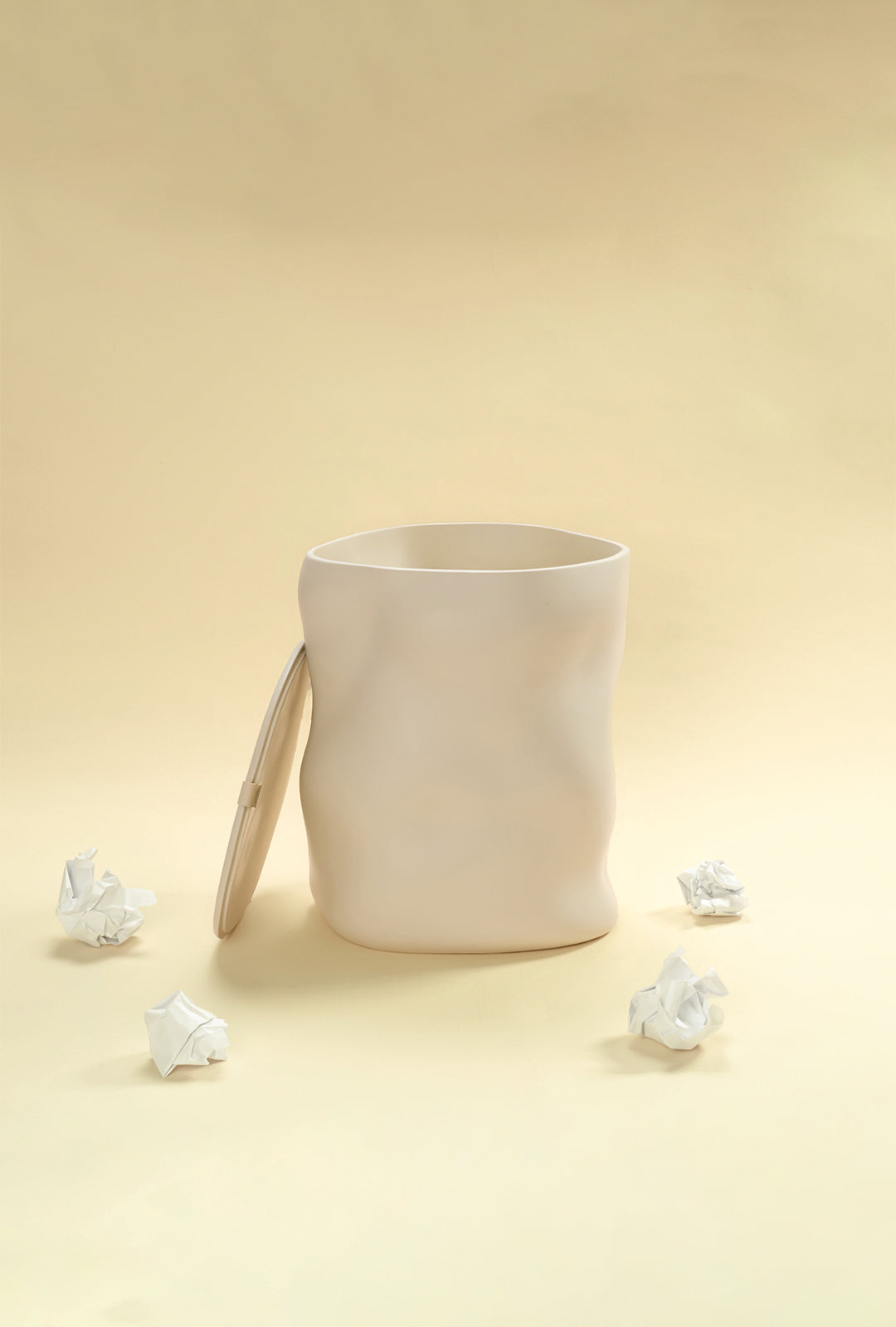 A beige sculptural dustbin shown open with its lid leaning against the side, surrounded by several crumpled pieces of white paper, against a light yellow background.