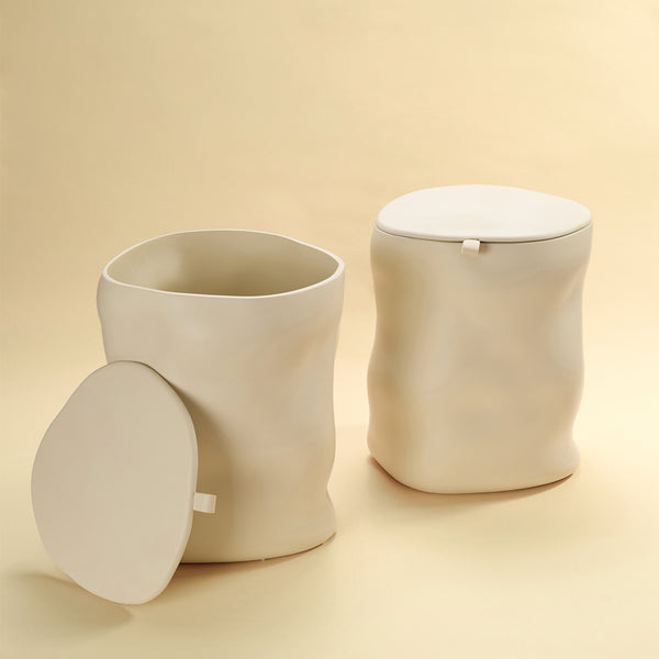 Two beige sculptural dustbins displayed side by side, one shown open with its lid placed to the side and the other shown closed, against a light yellow background.