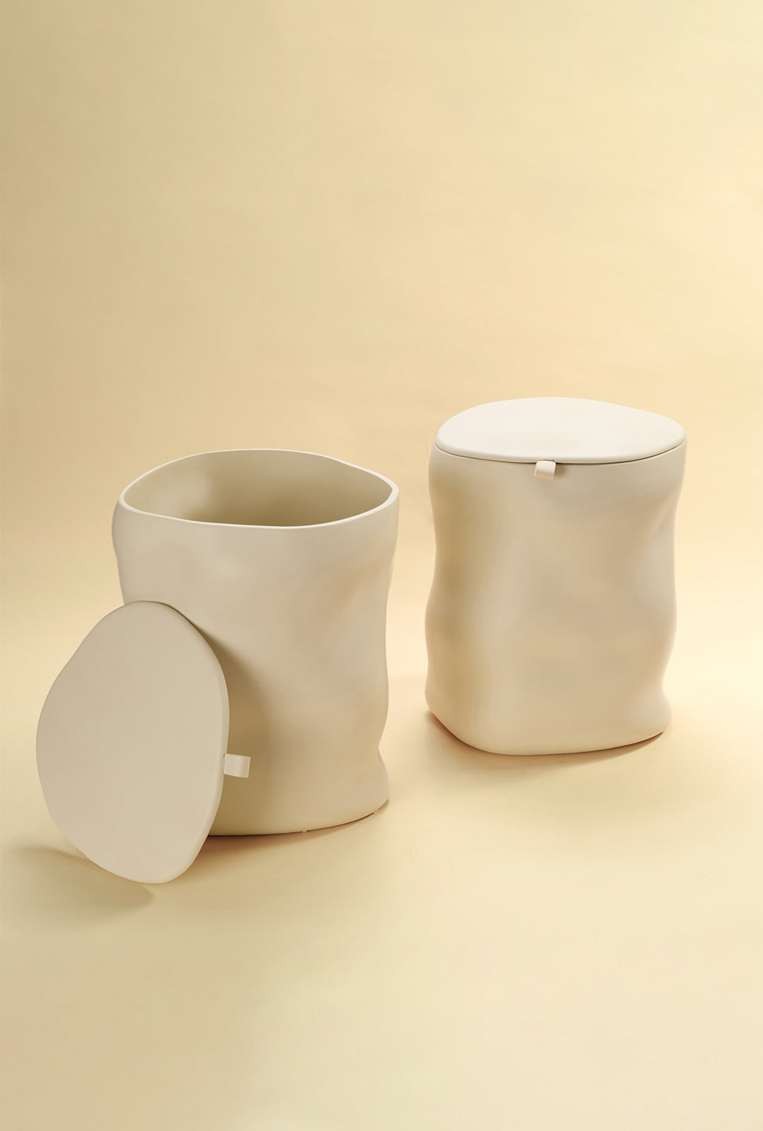 Two beige sculptural dustbins displayed side by side, one shown open with its lid placed to the side and the other shown closed, against a light yellow background.