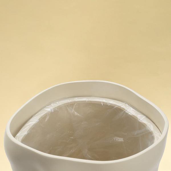 A close-up view of the open top of a beige sculptural dustbin, showing a clear plastic liner fitted inside, against a light yellow background.