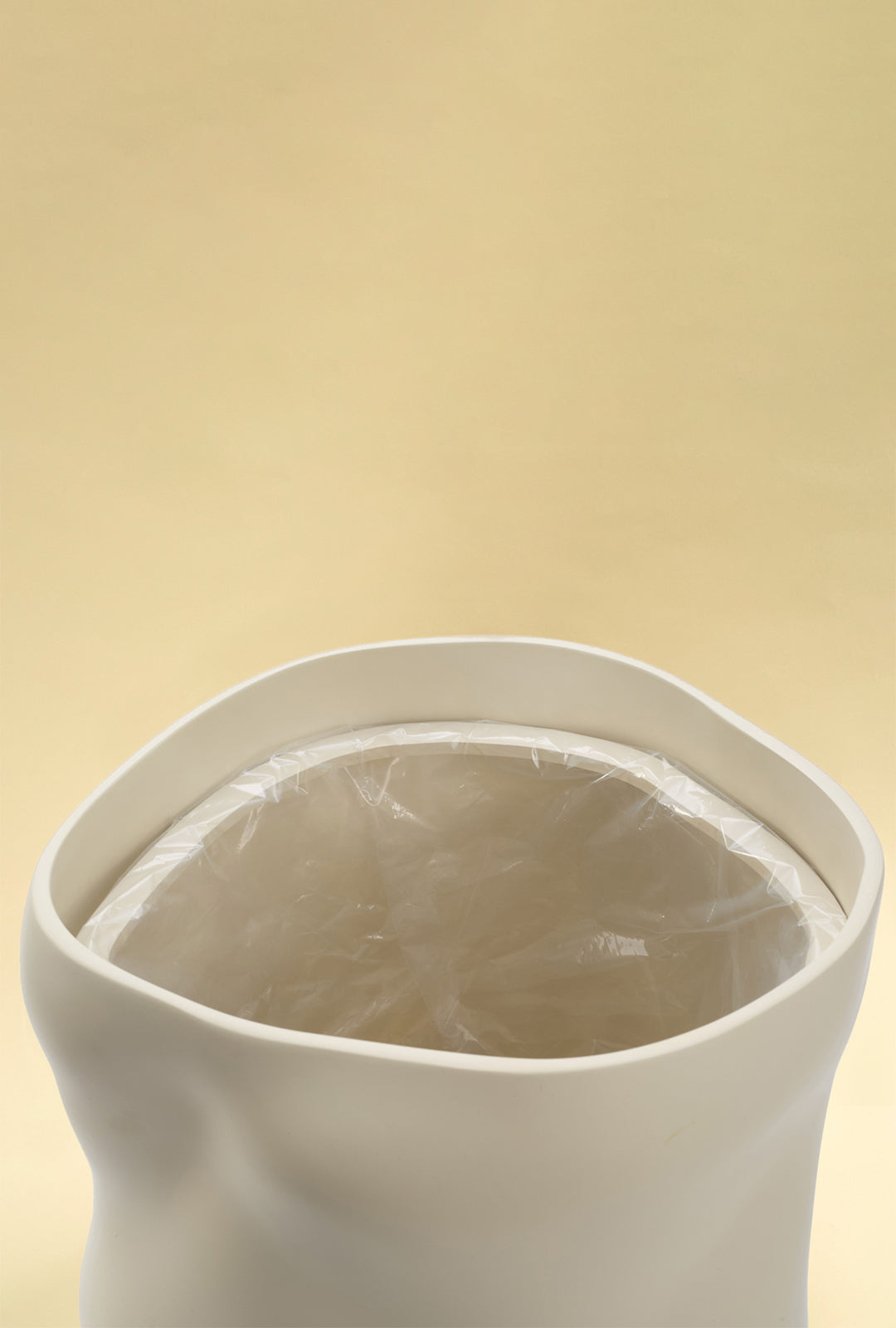 A close-up view of the open top of a beige sculptural dustbin, showing a clear plastic liner fitted inside, against a light yellow background.