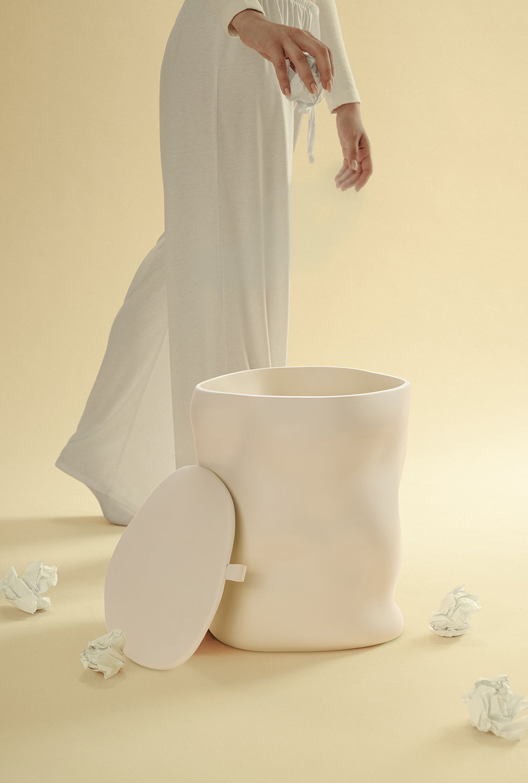 A beige sculptural dustbin shown with its lid placed to the side, as a person drops a crumpled piece of white paper into the open bin, against a light yellow background.