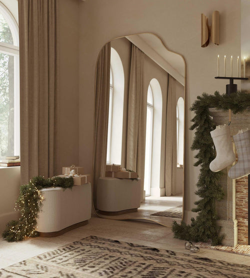 Asymmetrical full-length mirror with an organic shape and thin brass-colored frame, styled in a softly lit living space with holiday decor and arched windows.