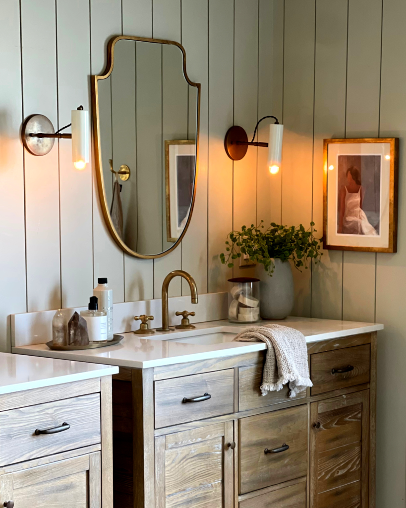 Shield-shaped wall mirror with a gold frame, mounted vertically above a bathroom vanity between two wall sconces.