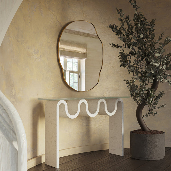 Irregular-shaped wall mirror with a thin brass-colored frame mounted above a sculptural console table in a textured interior setting.
