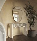 Irregular-shaped wall mirror with a thin brass-colored frame mounted above a sculptural console table in a textured interior setting.